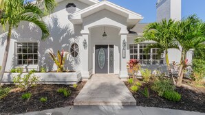 A white house with a central entrance, a small porch, and a garden with palm trees and plants. - Newly Updated!  RiverView Retreat  Historic Fort Myers, FL 10 guests, Spa & pool (Fort Myers)