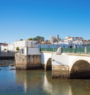 Eine Steinbrücke über einem ruhigen Fluss, im Hintergrund sind Gebäude zu sehen.