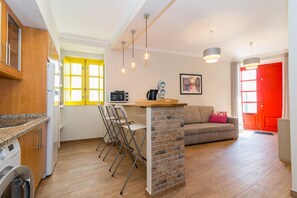 Eine moderne Küche mit integriertem Waschmaschinenanschluss, ein Essbereich mit Ziegelwand und ein Wohnbereich mit Sofa und roter Tür.