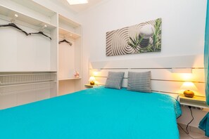 Ein Schlafzimmer mit einem Bett, zwei Nachttischlampen, einem Wandbild und einem Kleiderständer mit Kleidung.