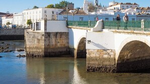 A stone bridge with arches over a calm river, leading to a town with white buildings and a clock tower. - Pegada Apartamento T3 Luxo (Tavira)