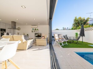 A modern living room with a sofa, chairs, a TV, and a kitchen area. - V3 Moradia Golden Nelly com Piscina (Tavira)