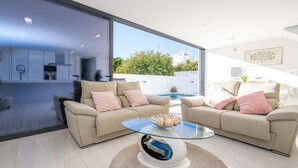 Ein modernes Wohnzimmer mit einem Glastisch, beigefarbenen Sofas und Blick auf einen Swimmingpool und Grünflächen.