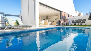 Ein moderner Poolbereich mit Liegestühlen, einem klaren blauen Pool und einem weißen Gebäude mit offener Tür zu einem Essbereich.