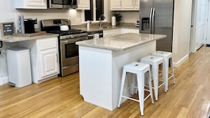 A modern kitchen with white cabinetry, stainless steel appliances, a granite countertop, and a central island with bar stools. - BOOKING for 2027! Walk to Town Neck Beach! LINENS-AC-New Amenities (Sandwich)