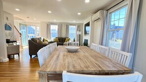 A spacious dining area with a long wooden table and white chairs, a cozy living room in the background, and large windows with curtains. - BOOKING for 2027! Walk to Town Neck Beach! LINENS-AC-New Amenities (Sandwich)