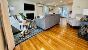 A spacious living room with a grey sofa, a small round table, and a TV mounted on the wall. - BOOKING for 2027! Walk to Town Neck Beach! LINENS-AC-New Amenities (Sandwich)