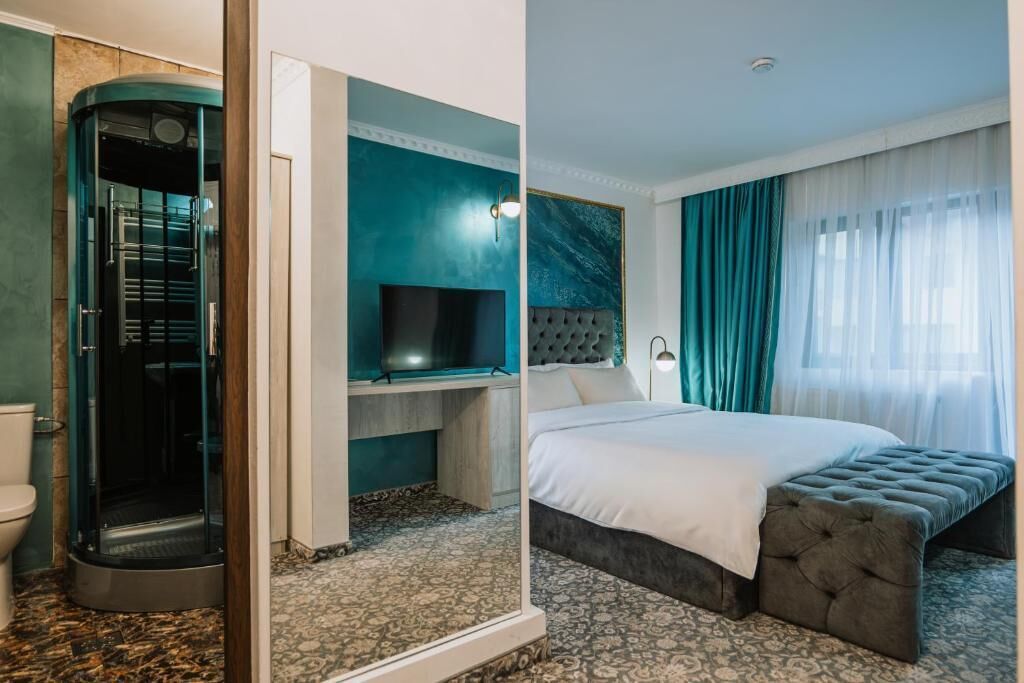 A hotel room with a curved shower enclosure, a flat-screen TV, a bed with a tufted headboard, and a large window with sheer curtains.