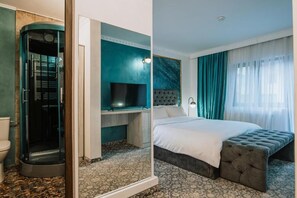 Habitación de hotel con cabina de ducha curva, televisor de pantalla plana, cama con cabecero capitonado y un amplio ventanal con cortinas translúcidas.