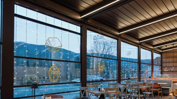 A modern cafe with large windows, wooden furniture, and a view of a cityscape. - Suru Hotel & Restaurant (Busteni)
