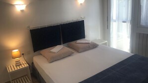 Ein ordentlich bezogenes Bett mit zwei Kissen und einer zusammengefalteten Decke, eine Nachttischlampe, ein kleiner weißer Nachttisch und ein an der Wand befestigter Bilderrahmen.