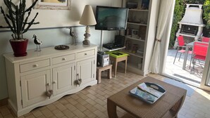Ein Wohnzimmer mit einem weißen Schrank, einem Fernseher, einem kleinen Tisch mit Zeitschriften, einer Topfpflanze und Blick ins Freie.