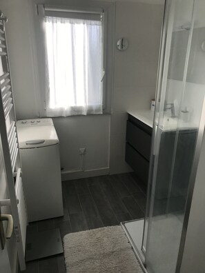 Een compacte badkamer met een wasmachine, een klein raam met dunne gordijnen, een spiegel en een douchehoek.