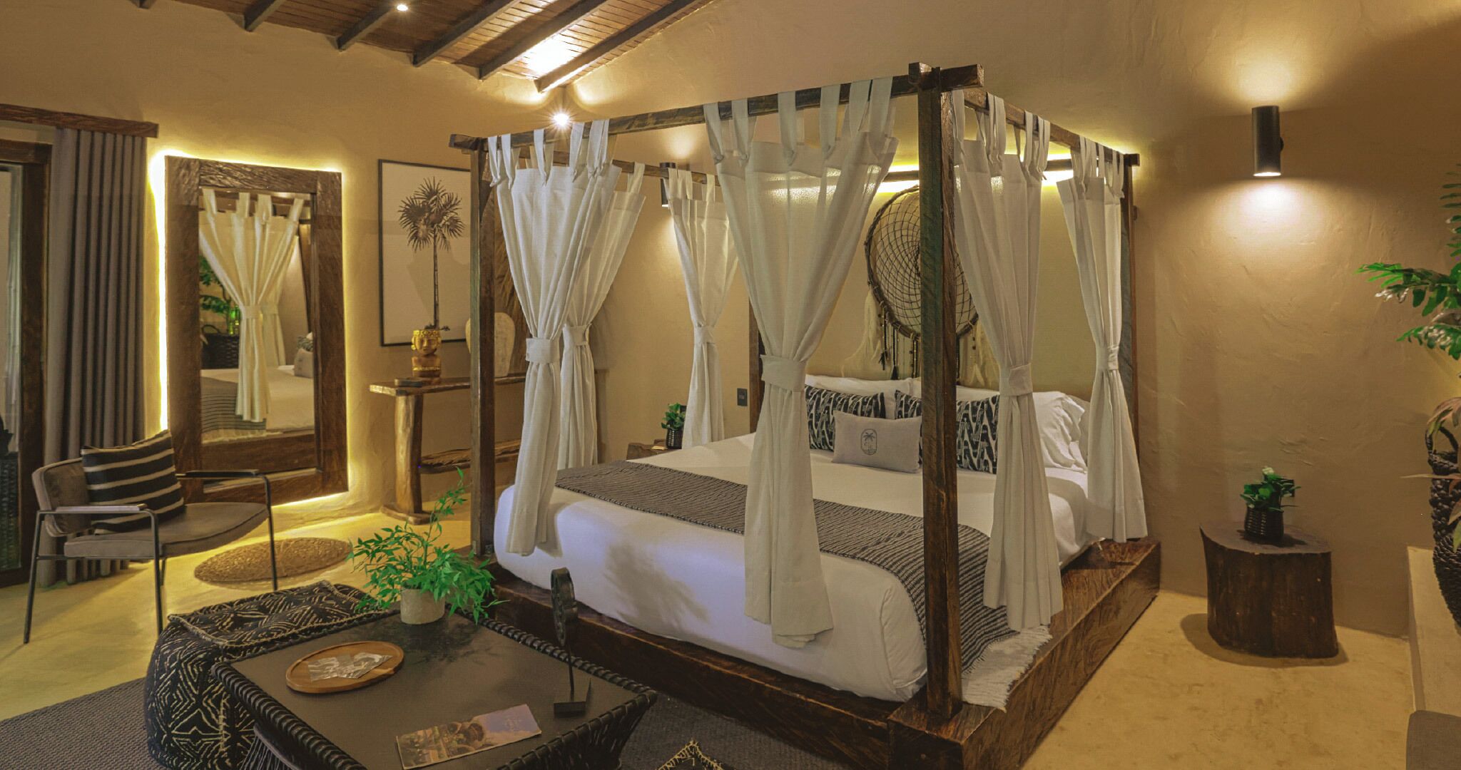 A four-poster canopy bed with white linens, a small side table, and a chair in a well-lit room with a wooden ceiling.