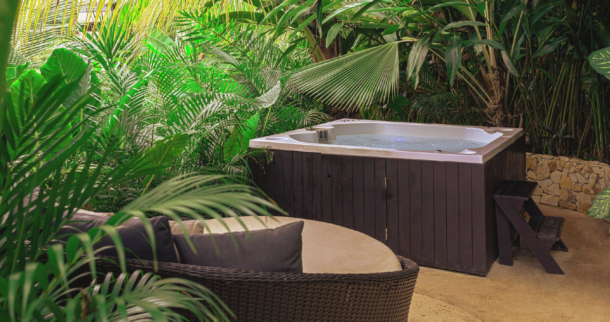 A hot tub surrounded by lush greenery, with a wicker chair and a stone wall nearby.
