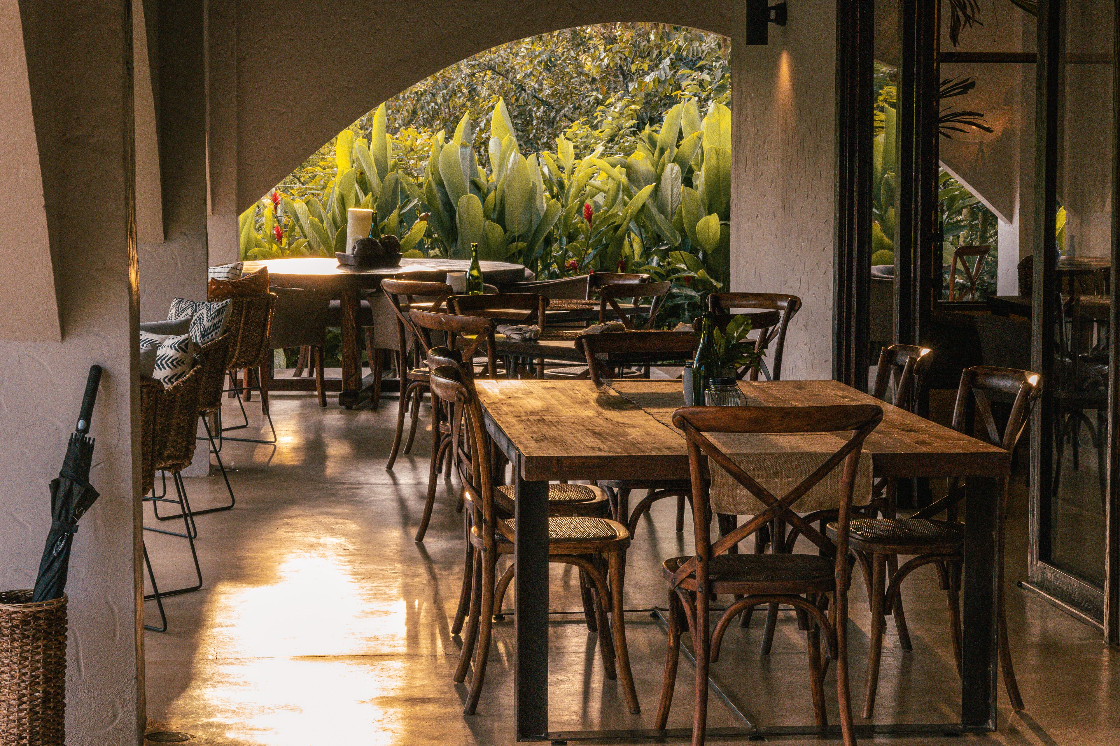 A restaurant with wooden tables and chairs, a large window with a view of plants, and a candle on the table.