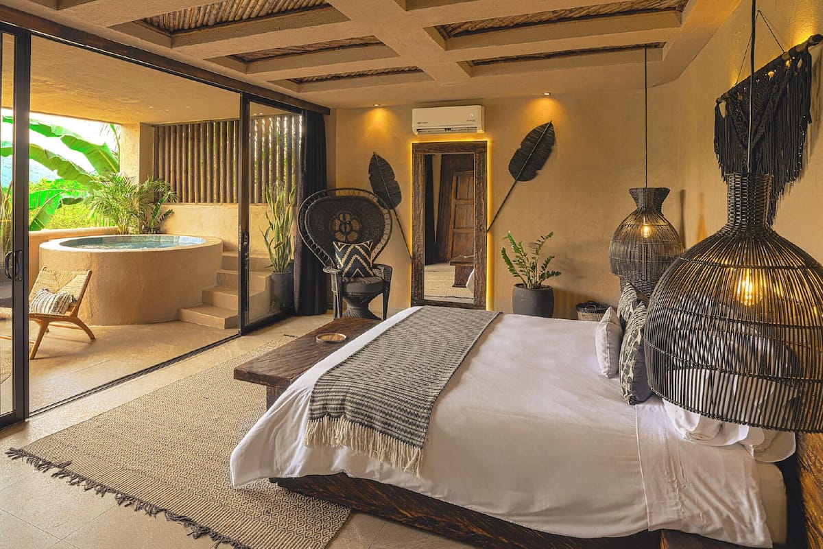 Une chambre spacieuse avec un grand lit, une porte vitrée donnant sur un balcon équipé d’un coin salon extérieur et d’un jacuzzi.