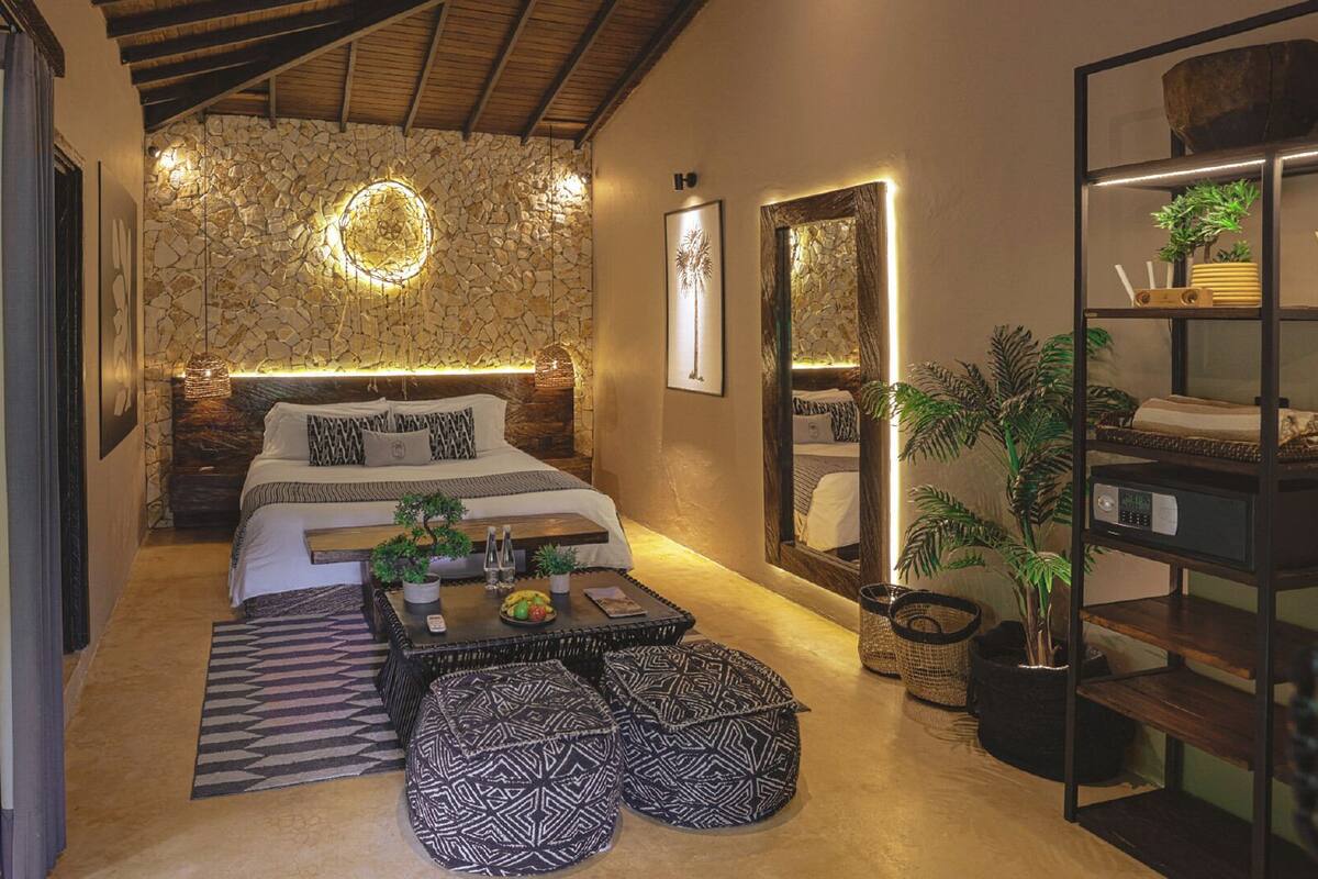 Une chambre avec un mur en pierre, un lit avec une tête de lit, une table de chevet avec une lampe, un miroir et une étagère avec des plantes.