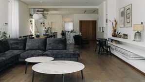 Een moderne woonkamer met een hoekbank, twee ronde salontafels, een eethoek met tafel en stoelen, en een keuken op de achtergrond.