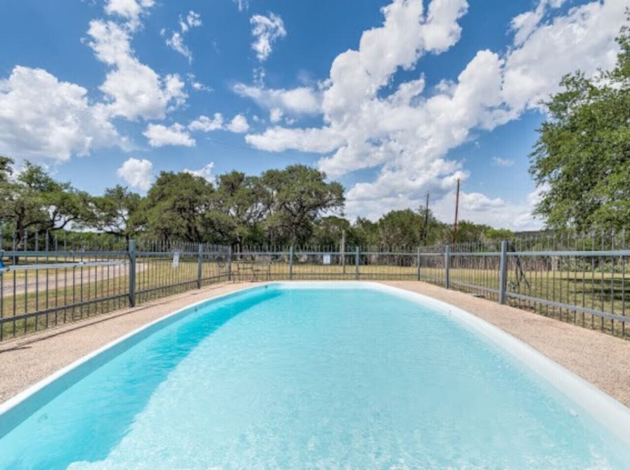 Uma piscina retangular com superfície de água azul e límpida, cercada por uma grade de metal e árvores ao fundo.
