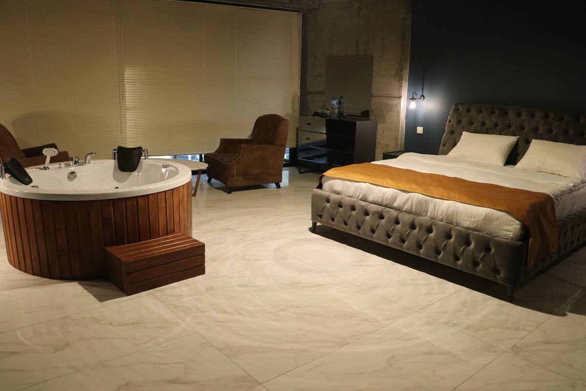 A bedroom with a large bed, a sitting area with a chair, a bathtub, and a wooden bench.