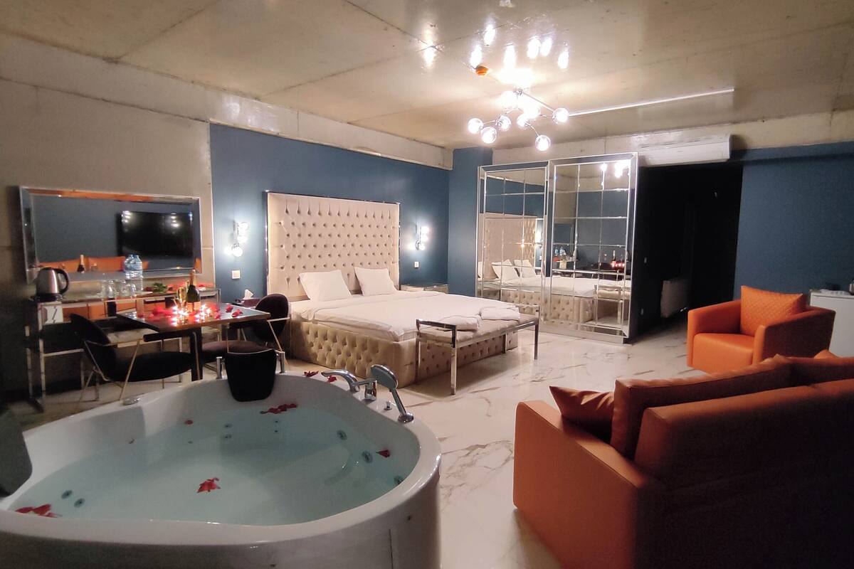A bedroom with a large bed, a sitting area with a sofa, a bathtub, and a dining area with a table and chairs.