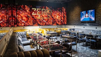 A modern lounge area with a large digital screen displaying 'ZERO ZERO' by Mayson, plush seating, and a variety of tables and chairs.