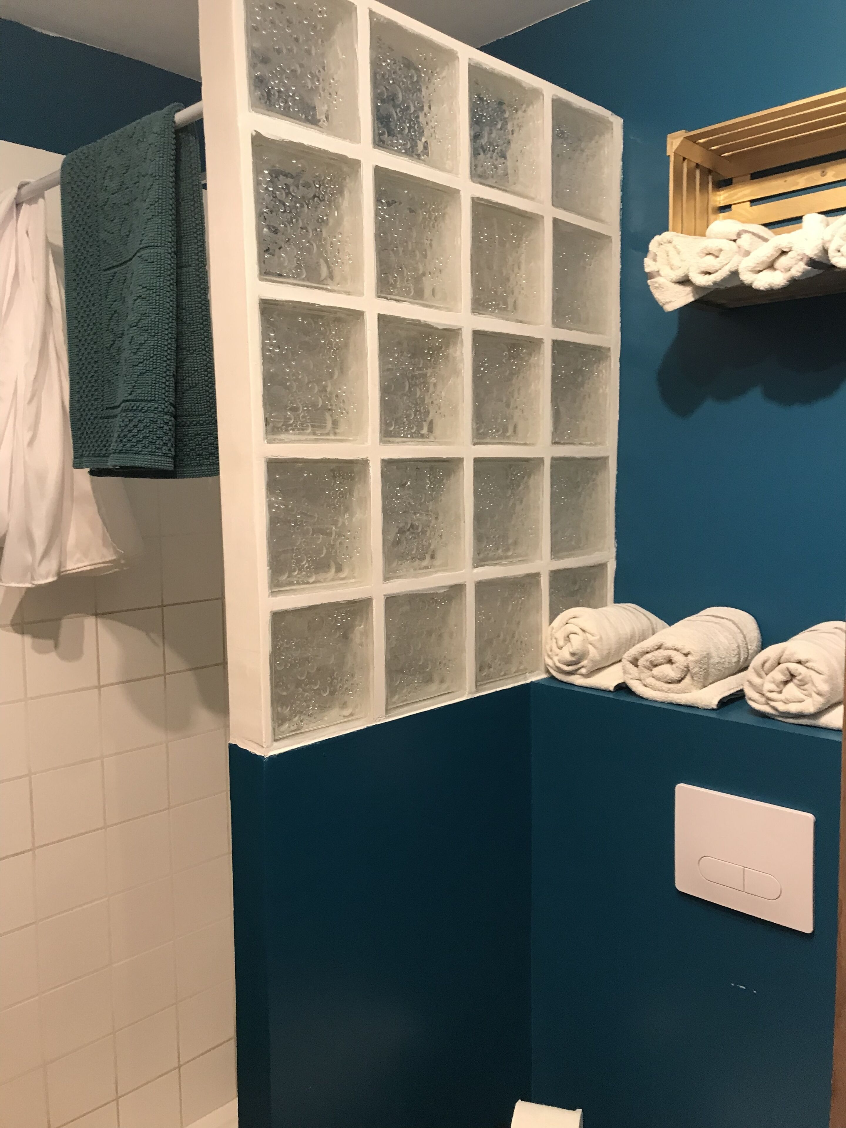 A bathroom with a blue wall, a white tiled shower area, a frosted glass partition, and a shelf with rolled towels.