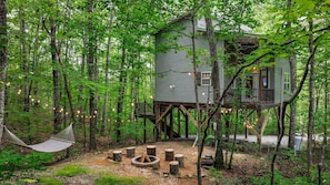 Une cabane dans les arbres avec une terrasse et un foyer, le tout entouré d’arbres.