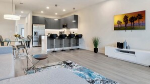 A modern kitchen with grey cabinets, a stainless steel refrigerator, and a microwave. There is a dining area with a glass table and chairs, and a living room with a sofa and a coffee table.