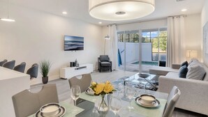 A modern living room with a dining area, a grey sofa, a large window, and a view of the outdoors.