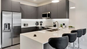 A modern kitchen with grey cabinets, stainless steel appliances, a central island with bar stools, and recessed lighting.