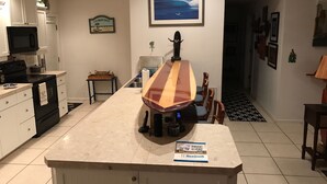 Una cocina con una isla grande, microondas y refrigerador. Hay un cuadro enmarcado en la pared y una mesita con una cesta.