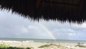 Vista de una playa con una estructura de techo de paja, un arcoíris visible y una playa arenosa con vegetación escasa.