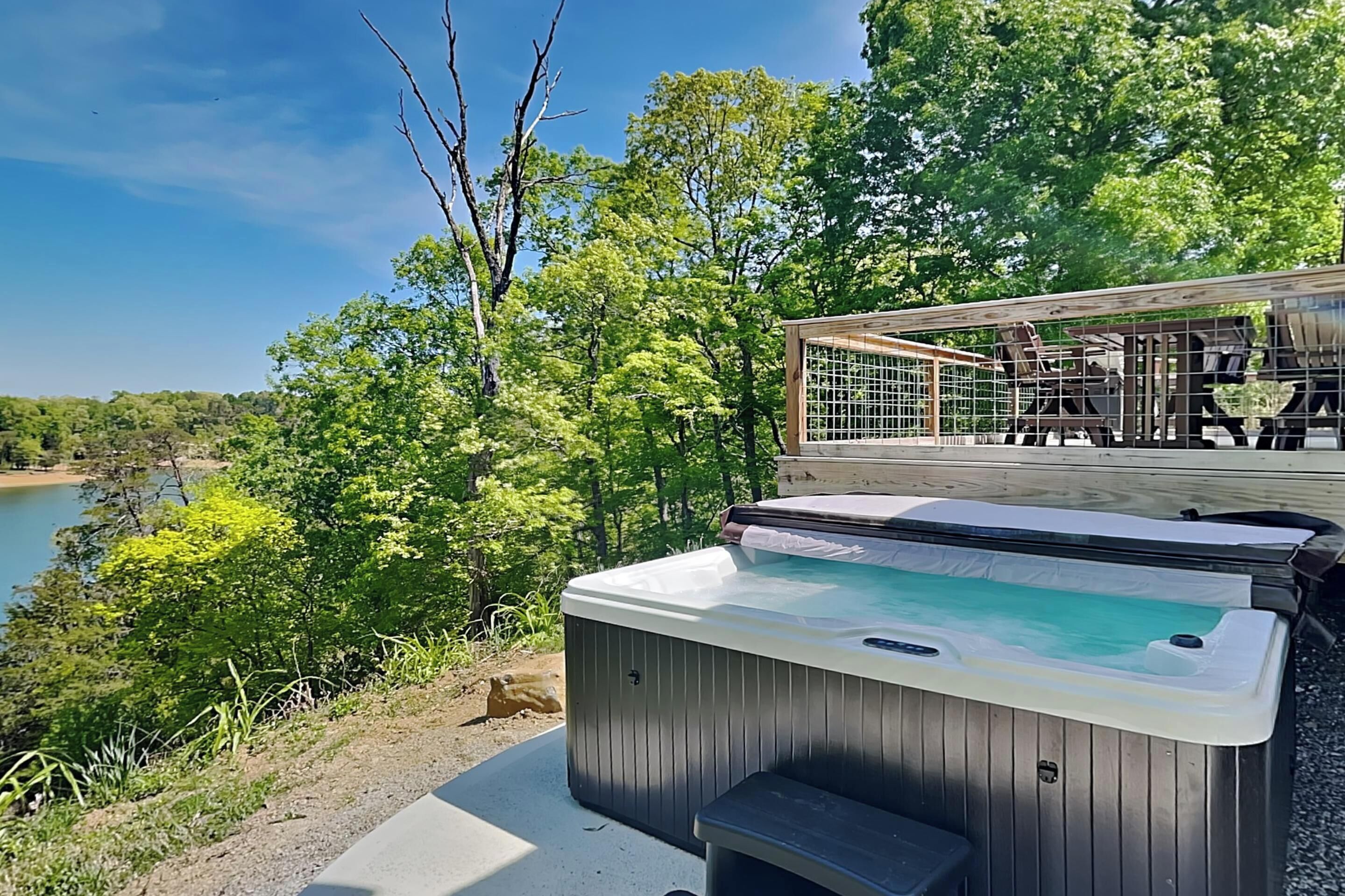 A hot tub with a view of a lake and trees, a deck with chairs, and a wooden fence.
