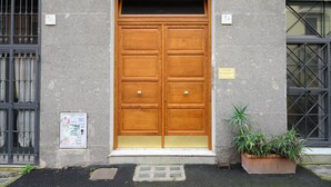 Eine hölzerne Doppeltür mit einem kleinen Fenster darüber, eine Topfpflanze rechts und ein Metallgitter links.