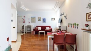 Ein Wohnzimmer mit einem roten Sofa, einem Esstisch mit Stühlen und einer Küchenzeile, inklusive Kühlschrank und Topfpflanzen.