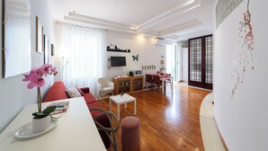 Ein modernes Wohnzimmer mit einer Couch, einem kleinen Tisch mit einer Pflanze, einem Fernseher und einer Küchenzeile im Hintergrund.