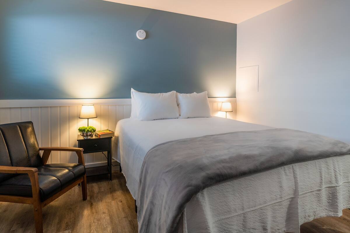 A neatly arranged bedroom with a bed, a chair, a bedside table with a lamp, and a wall-mounted light fixture.