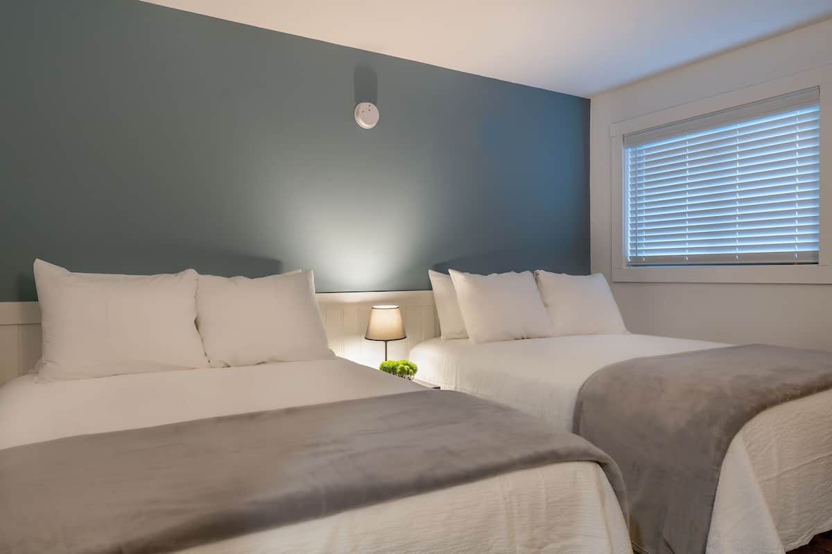 A hotel room with two beds, a nightstand with a lamp, and a window with blinds.
