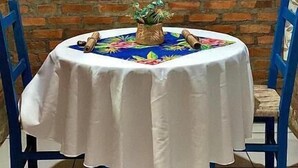 A round table with a white tablecloth, blue chairs, and a framed floral artwork on the brick wall behind. - Pousada & Camping Nativos dos Canyons (Praia Grande)