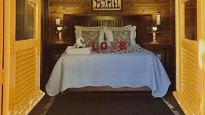 A cozy room with wooden walls and a sloped ceiling, featuring a bed with a white bedspread and decorative pillows, a framed artwork above the bed, and a small table with a lamp. - Pousada & Camping Nativos dos Canyons (Praia Grande)