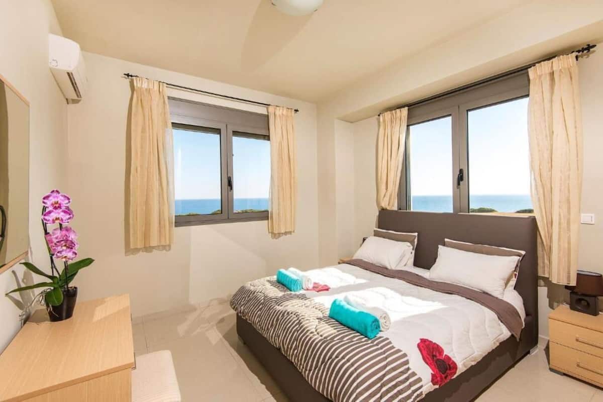 A hotel room with a large bed, two windows with curtains, a bedside table, a desk, and a potted orchid.