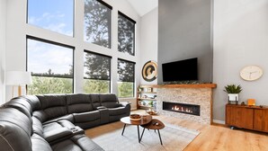 A modern living room with a sectional sofa, a fireplace, a flat-screen TV, and large windows. - Copper Skies Lodge In Powder House Pass (South Lawrence)