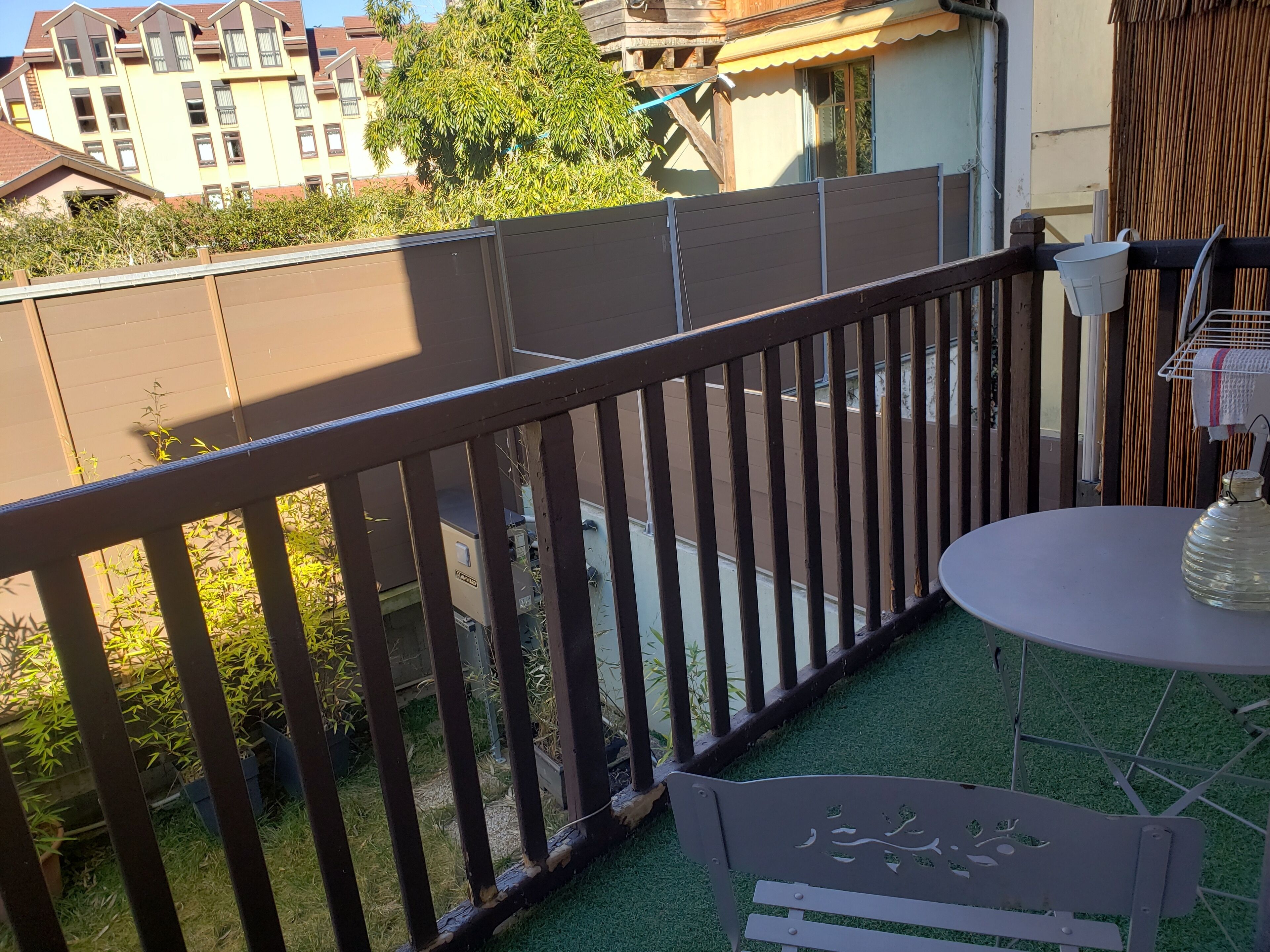 Ein Balkon mit Metallgeländer, einem kleinen runden Tisch und einer Flasche. Im Hintergrund sind Gebäude und ein Baum zu sehen.
