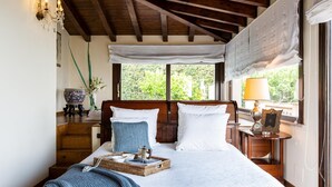 Quarto com cabeceira de madeira, cama com roupa de cama branca, bandeja de vime com livros, abajur e vista para o verde lá fora.