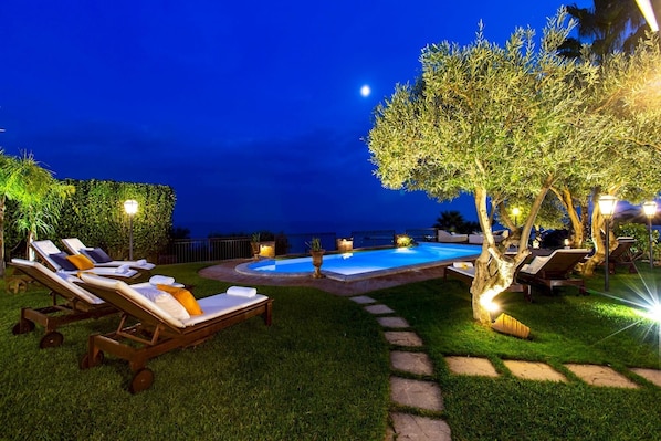 A nighttime outdoor pool area with lounge chairs and a tree.