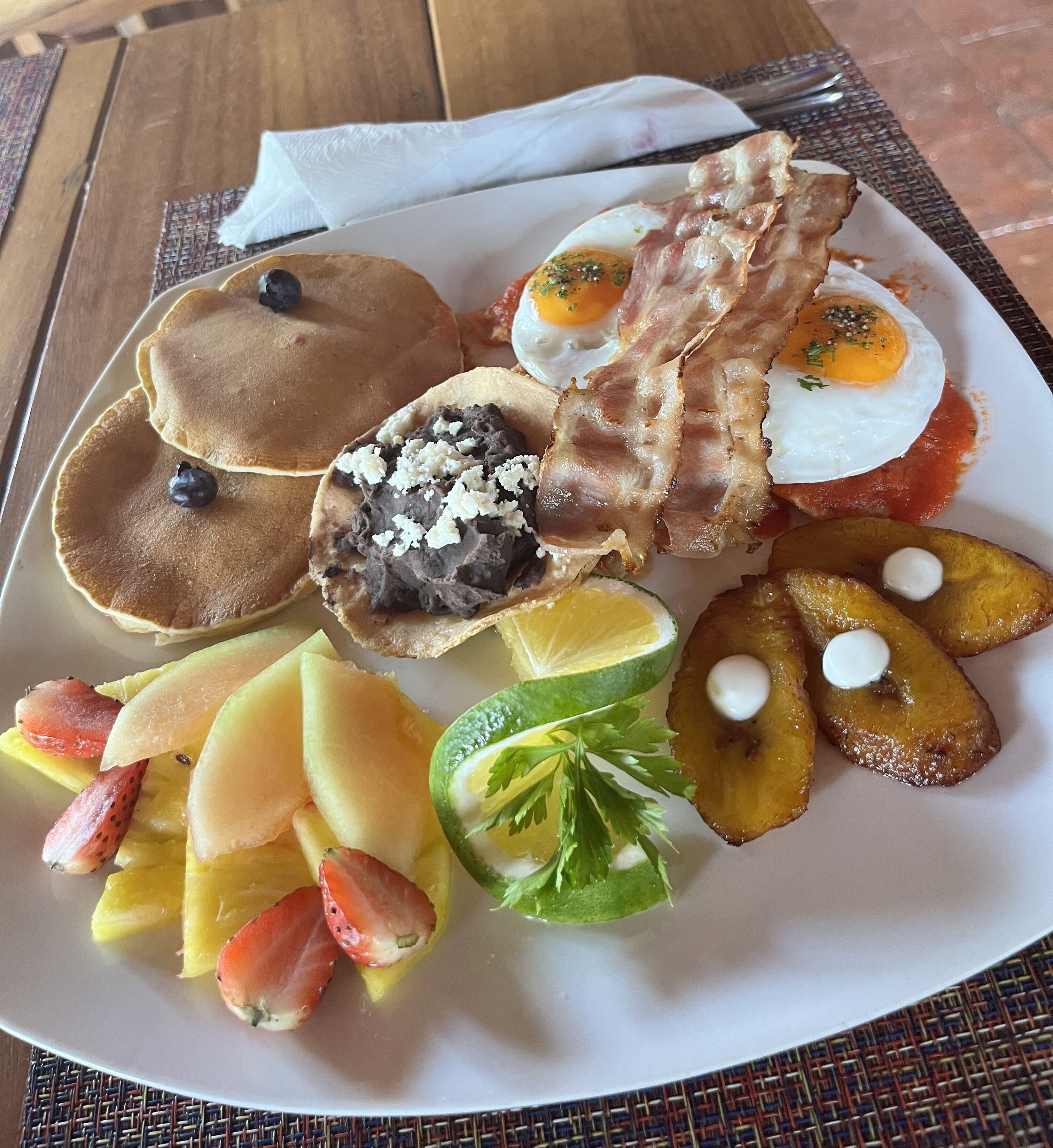 Een ontbijtbord met pannenkoeken, spek, eieren en fruit.