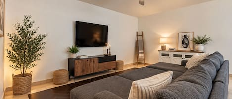 A modern living room with a large grey sofa, a flat-screen TV, a ceiling fan, and a sideboard with decorative items.