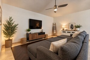 A modern living room with a large grey sofa, a flat-screen TV, a ceiling fan, and a sideboard with decorative items. - Stunning 7bed Oasis-Pickleball,Heated Pool, hot tub, waterslide (Scottsdale)